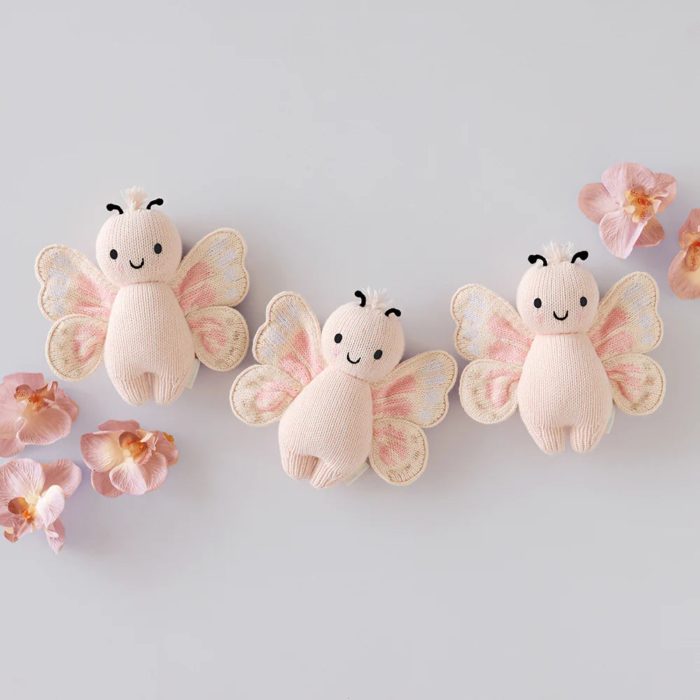 Three Cuddle + Kind Baby Butterfly - Petal Pink plush toys are displayed on a light surface with pink artificial flowers, showcasing their collectible, heirloom-quality charm.