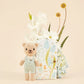Tiny Charlie the Honey Bear by Cuddle + Kind stands beside a floral gift box, with white and yellow flowers arranged behind it.