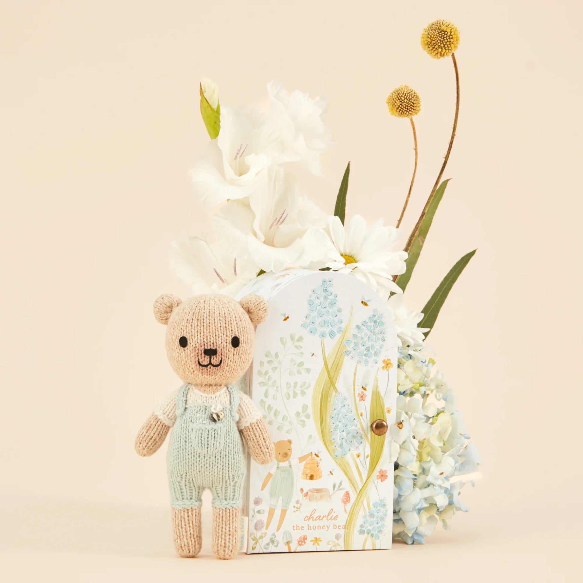 Tiny Charlie the Honey Bear by Cuddle + Kind stands beside a floral gift box, with white and yellow flowers arranged behind it.