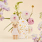 Tiny Olivia the Honey Bear by Cuddle + Kind stands in a white dress before a floral box, surrounded by pastel flowers—a charming, artisan-made fair trade doll perfect for any collection.