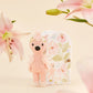 Tiny Penelope the Flamingo, a hand-knit doll by Cuddle + Kind, stands before a floral-patterned box, surrounded by pink lilies on a light background.