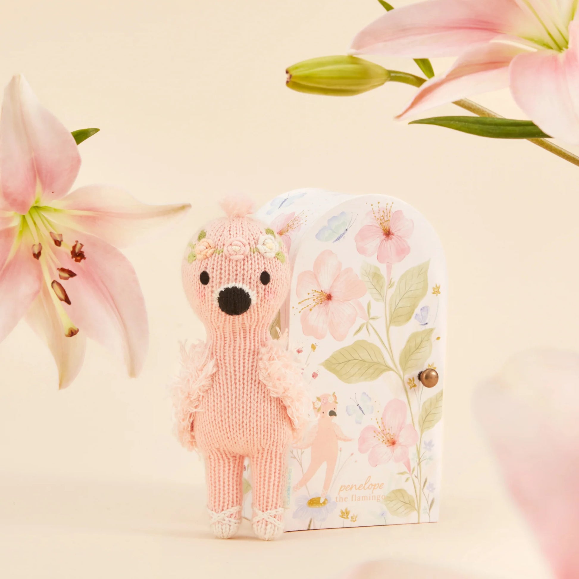 Tiny Penelope the Flamingo, a hand-knit doll by Cuddle + Kind, stands before a floral-patterned box, surrounded by pink lilies on a light background.