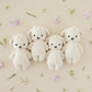 Four Cuddle + Kind Baby Lamb plush toys, cream-colored and heirloom-quality, are lined up on a beige surface with scattered small flowers and greenery.