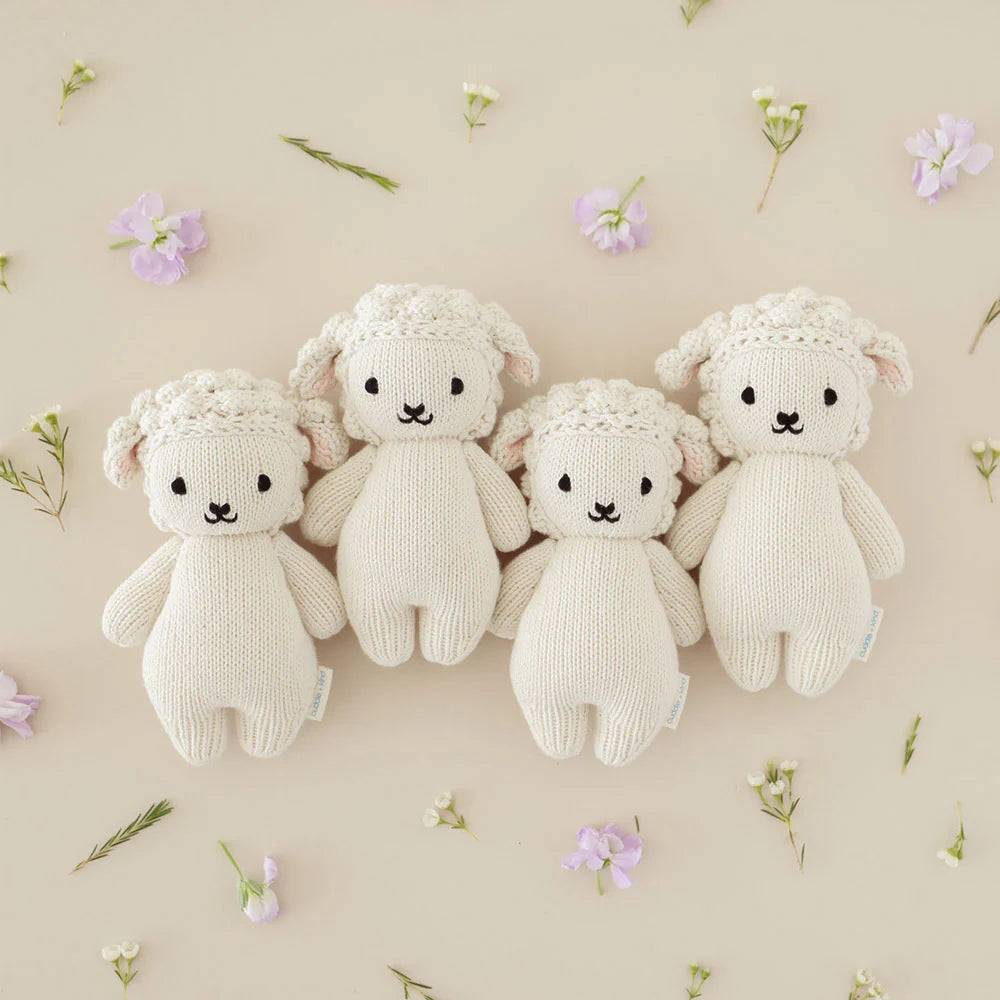 Four Cuddle + Kind Baby Lamb plush toys, cream-colored and heirloom-quality, are lined up on a beige surface with scattered small flowers and greenery.