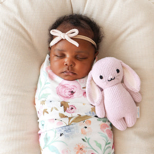 A baby wearing a floral swaddle and a pink headband sleeps while holding the Cuddle + Kind Lilac Bunny - Baby 7", lovingly hand-knit from the softest Peruvian cotton yarn, on a light textured surface.