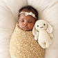 A baby wrapped in a yellow floral blanket sleeps on a cushioned surface next to the Cuddle + Kind Oatmeal Bunny - Baby 7", an heirloom-quality, cream-colored stuffed bunny toy.