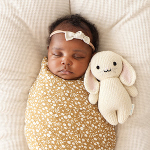 A baby wrapped in a yellow floral blanket sleeps on a cushioned surface next to the Cuddle + Kind Oatmeal Bunny - Baby 7", an heirloom-quality, cream-colored stuffed bunny toy.