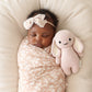 A baby wrapped in a pink floral blanket, wearing a pink headband, sleeps next to the Cuddle + Kind "Rose Bunny - Baby 7"," an heirloom-quality, hand-knit baby bunny made from soft Peruvian cotton yarn, on a cream-colored cushion.
