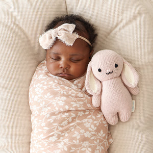 A baby wrapped in a pink floral blanket, wearing a pink headband, sleeps next to the Cuddle + Kind "Rose Bunny - Baby 7"," an heirloom-quality, hand-knit baby bunny made from soft Peruvian cotton yarn, on a cream-colored cushion.