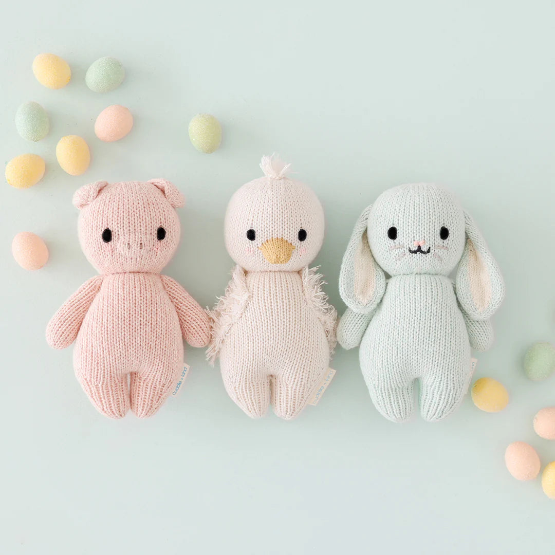Cuddle + Kind’s hand-knit Baby Gosling, crafted from soft Peruvian cotton yarn, is displayed in a row with pastel eggs on a light background.