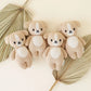 Four Cuddle + Kind “Baby Puppy” knitted tan plush dog toys, each with a white belly and face and handcrafted from soft cotton yarn, are arranged in a row on two dried palm leaves against a light-colored background.