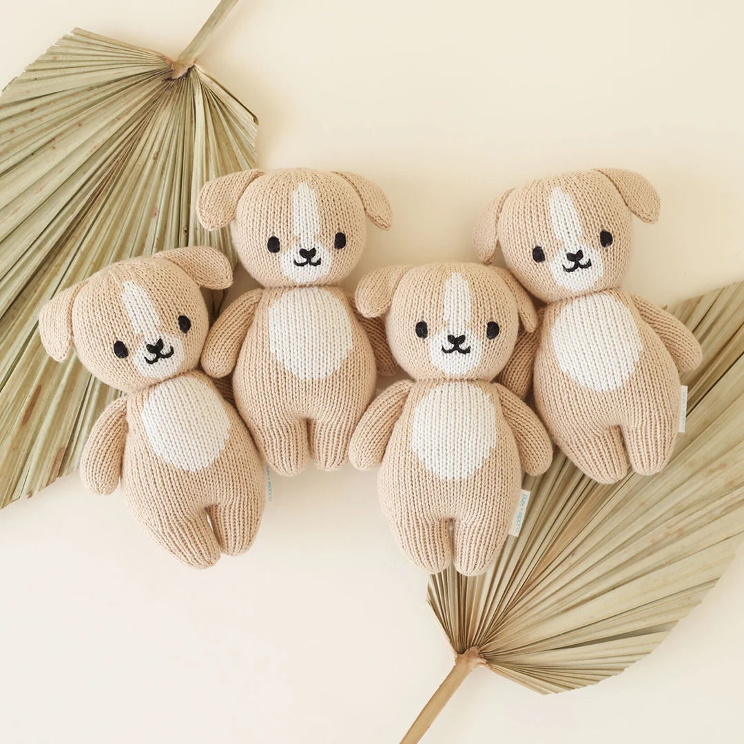 Four Cuddle + Kind “Baby Puppy” knitted tan plush dog toys, each with a white belly and face and handcrafted from soft cotton yarn, are arranged in a row on two dried palm leaves against a light-colored background.