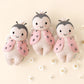 Three Cuddle + Kind Baby Ladybug plush dolls, each hand-knit with pink wings and smiling faces, are displayed side by side on a light background surrounded by small white flowers.