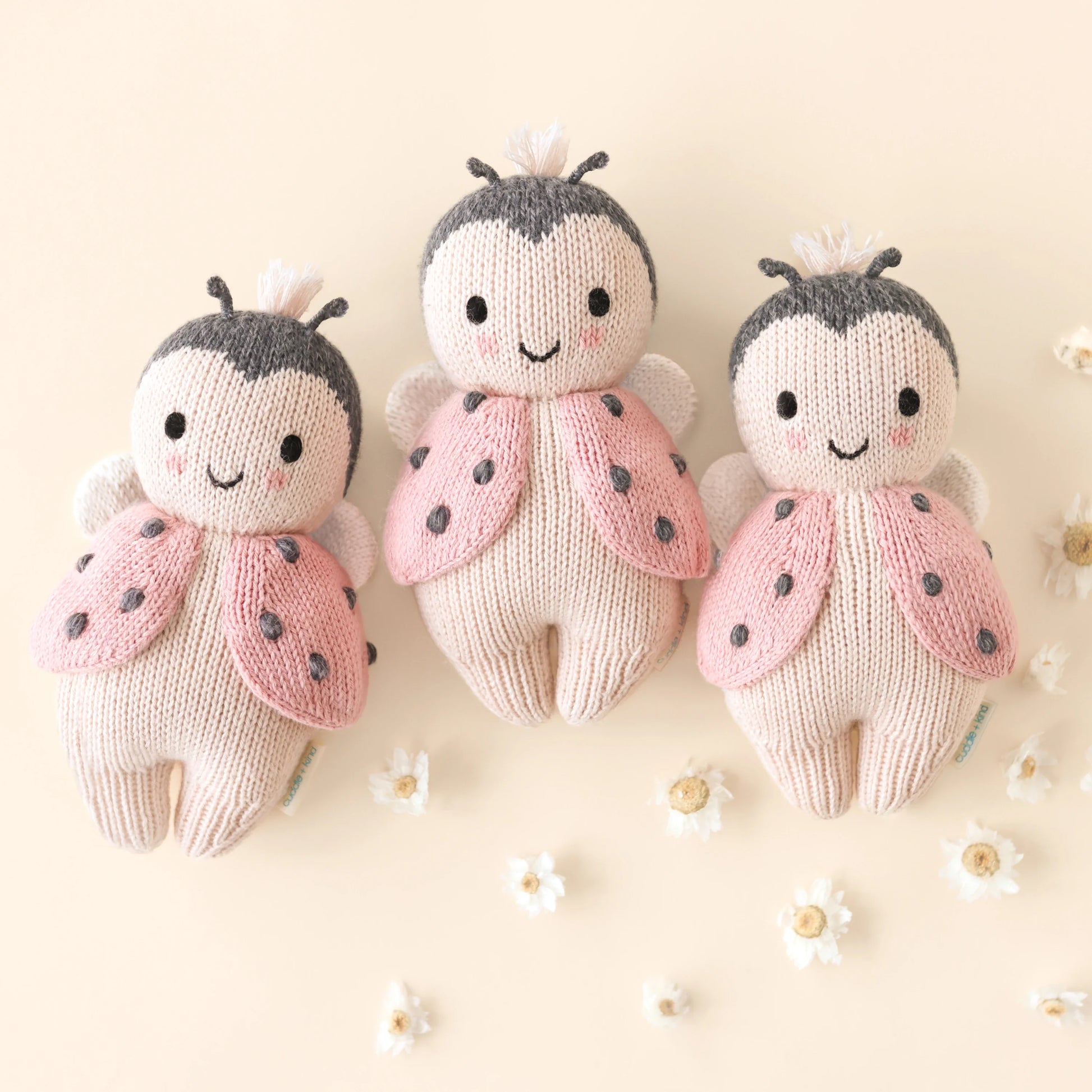 Three Cuddle + Kind Baby Ladybug plush dolls, each hand-knit with pink wings and smiling faces, are displayed side by side on a light background surrounded by small white flowers.