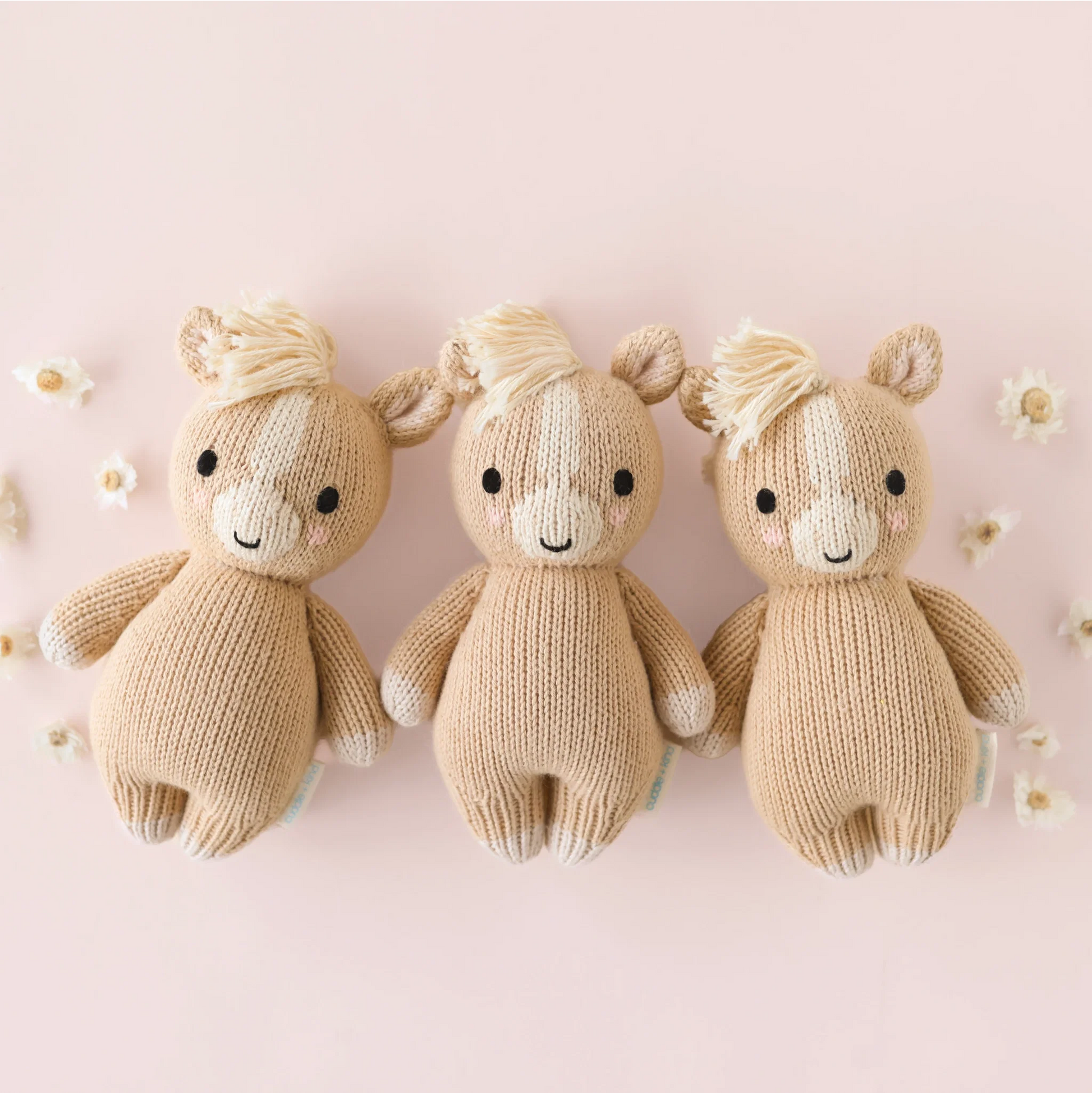 Three identical Baby Pony dolls by Cuddle + Kind—each a soft baby toy carefully handcrafted from cotton yarn—are arranged side by side against a light pink background adorned with scattered small white flowers.