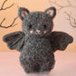 A cozy baby companion, the Cuddle + Kind Baby Bat - Charcoal features textured gray fur, black wings, and pink inner ears, shown against a pale background with bare tree branches.