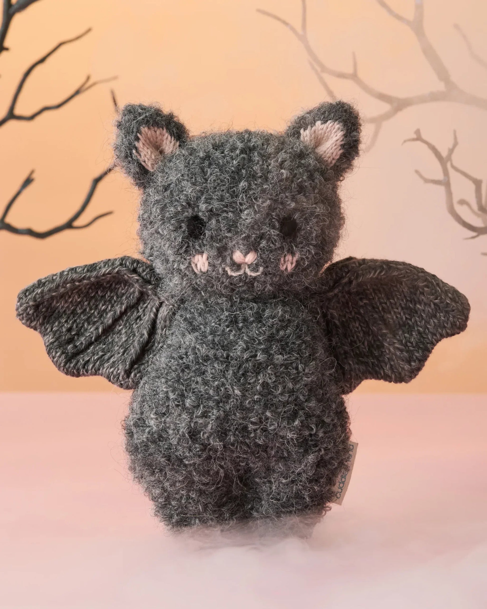 A cozy baby companion, the Cuddle + Kind Baby Bat - Charcoal features textured gray fur, black wings, and pink inner ears, shown against a pale background with bare tree branches.