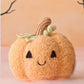 A handcrafted Baby Pumpkin by Cuddle + Kind, featuring a smiling face, embroidered eyes, and a green stem made from cotton yarn, sits atop a pink surface backed by a peach background with black branches. This charming heirloom plush is the perfect soft companion for cozy playtime or serves as a whimsical accent in any nursery.
