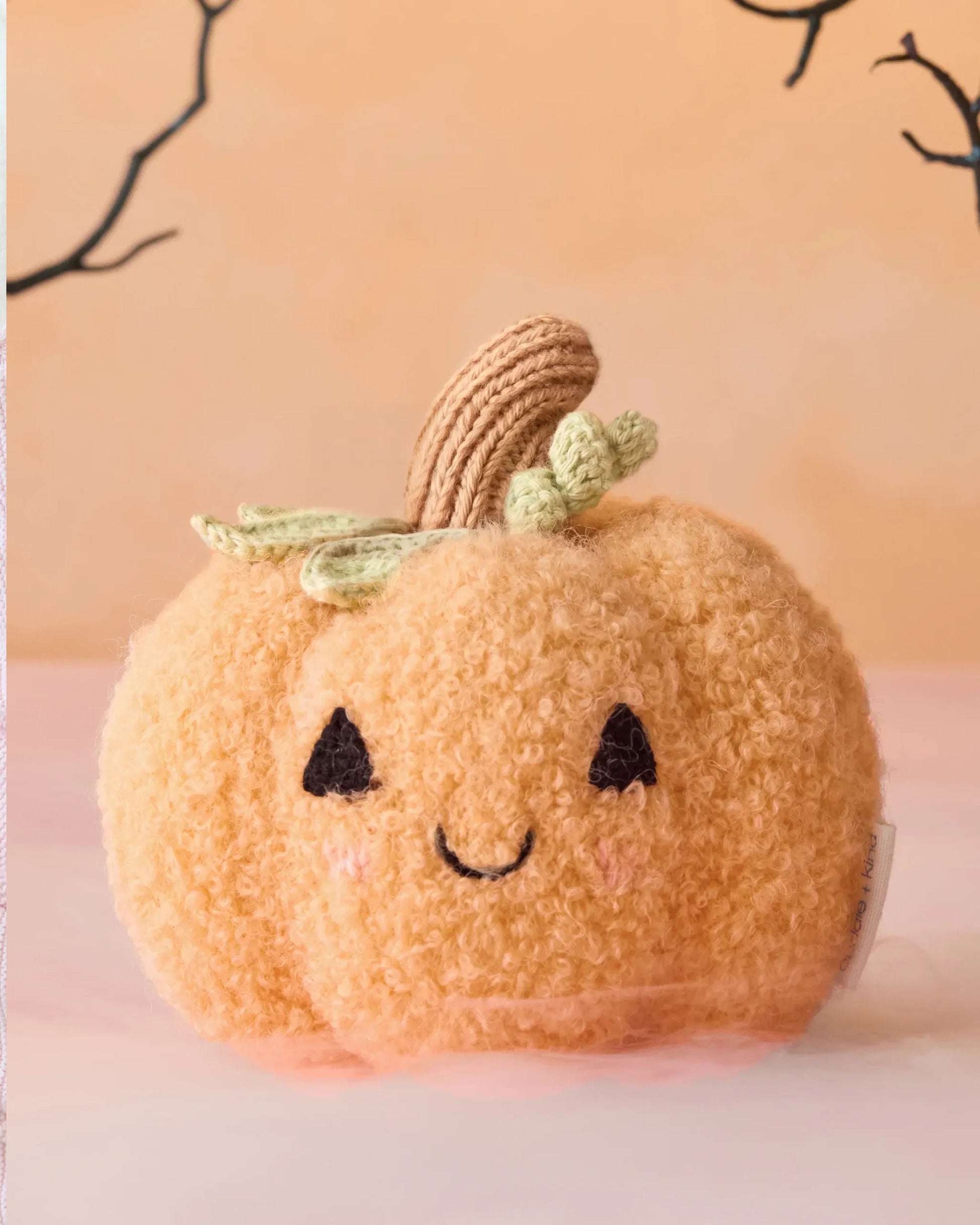 A handcrafted Baby Pumpkin by Cuddle + Kind, featuring a smiling face, embroidered eyes, and a green stem made from cotton yarn, sits atop a pink surface backed by a peach background with black branches. This charming heirloom plush is the perfect soft companion for cozy playtime or serves as a whimsical accent in any nursery.