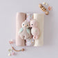 Three folded baby blankets and two Cuddle + Kind Baby Seahorse - Powder Pink plush toys, hand-knit from Peruvian cotton yarn, are surrounded by wooden and bead rattles plus small decor on a light surface.