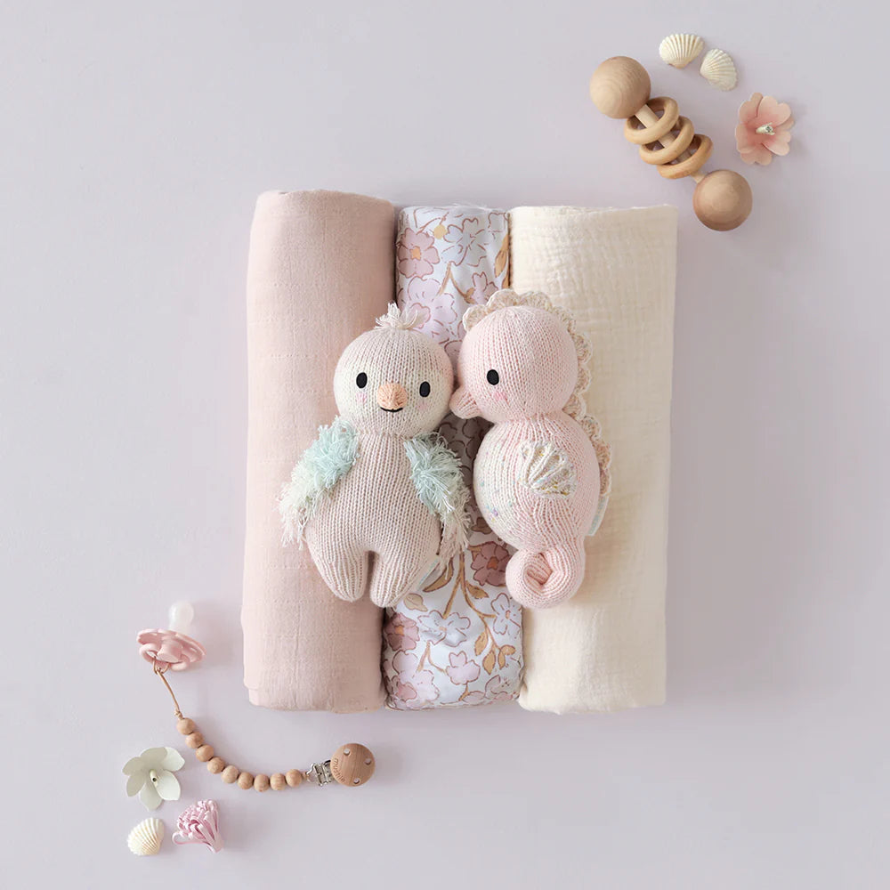 Three folded baby blankets and two Cuddle + Kind Baby Seahorse - Powder Pink plush toys, hand-knit from Peruvian cotton yarn, are surrounded by wooden and bead rattles plus small decor on a light surface.