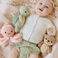 A baby lies on a blanket, wearing a white sweater and green checkered shorts, holding three hand-knit baby animals—each crafted from heirloom-quality Peruvian cotton yarn—including the Kelp Green Sea Turtle - Baby 7" by Cuddle + Kind, along with a pink octopus and a brown otter.