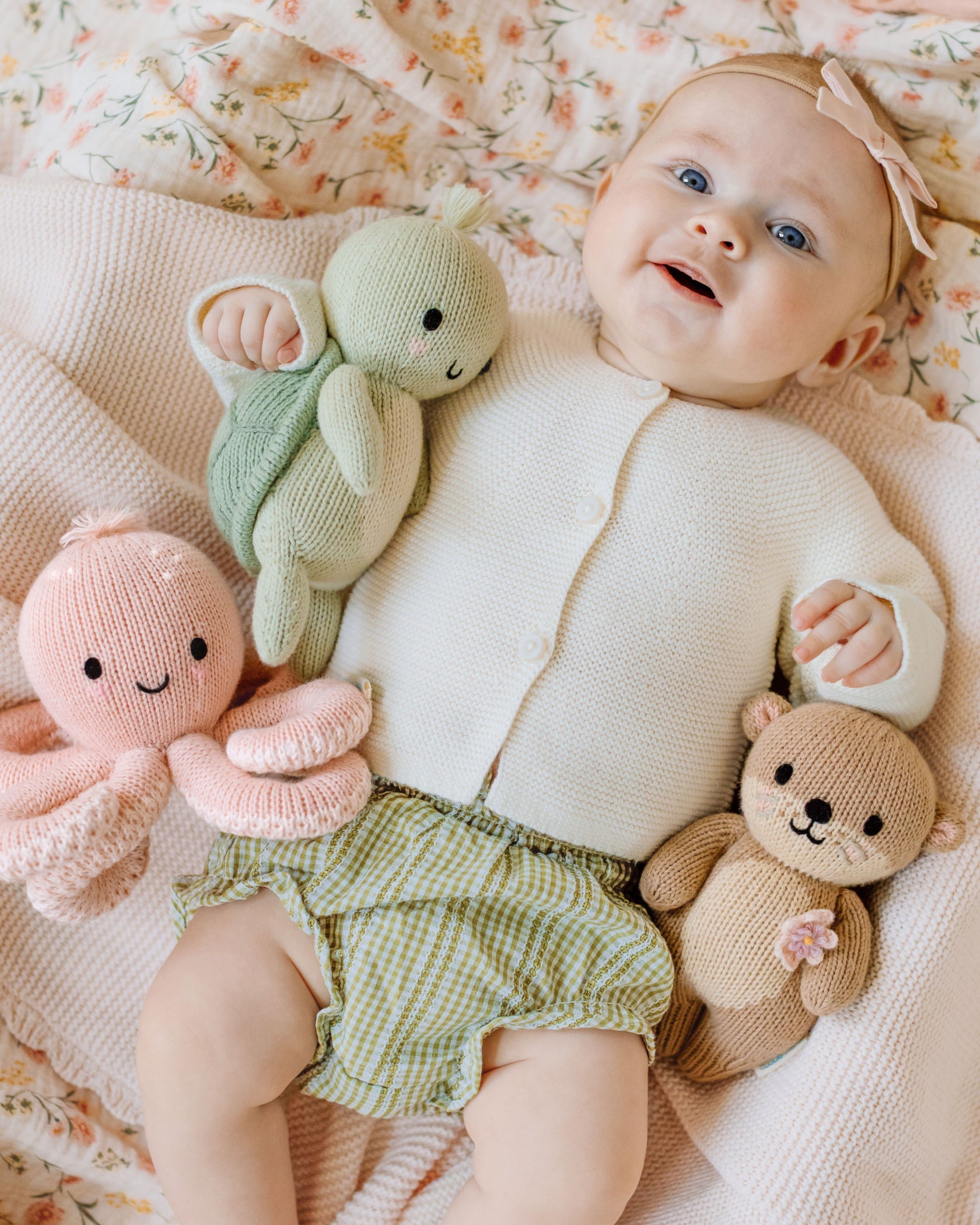 A baby lies on a blanket, wearing a white sweater and green checkered shorts, holding three hand-knit baby animals—each crafted from heirloom-quality Peruvian cotton yarn—including the Kelp Green Sea Turtle - Baby 7" by Cuddle + Kind, along with a pink octopus and a brown otter.