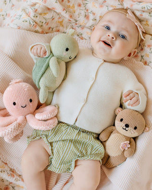 A baby lies on a blanket, wearing a white sweater and green checkered shorts, holding three hand-knit baby animals—each crafted from heirloom-quality Peruvian cotton yarn—including the Kelp Green Sea Turtle - Baby 7" by Cuddle + Kind, along with a pink octopus and a brown otter.