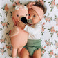 A baby with a pacifier, wearing a pink headband and green shorts, hugs Penelope the Flamingo Little 13" from Cuddle + Kind while lying on a floral-patterned blanket adorned with tropical flowers.