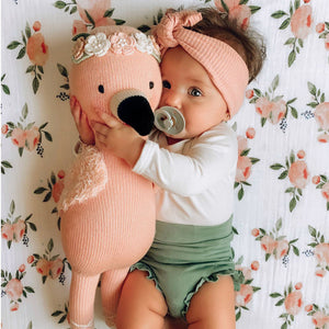 A baby with a pacifier, wearing a pink headband and green shorts, hugs Penelope the Flamingo Little 13" from Cuddle + Kind while lying on a floral-patterned blanket adorned with tropical flowers.