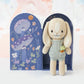 Meet Tiny Henry the Bunny—a hand-knit doll with long ears and a flower accessory from Cuddle + Kind's tiny collection, posed before an illustrated box of mushrooms and flowers. Ethically artisan-made and fair trade.