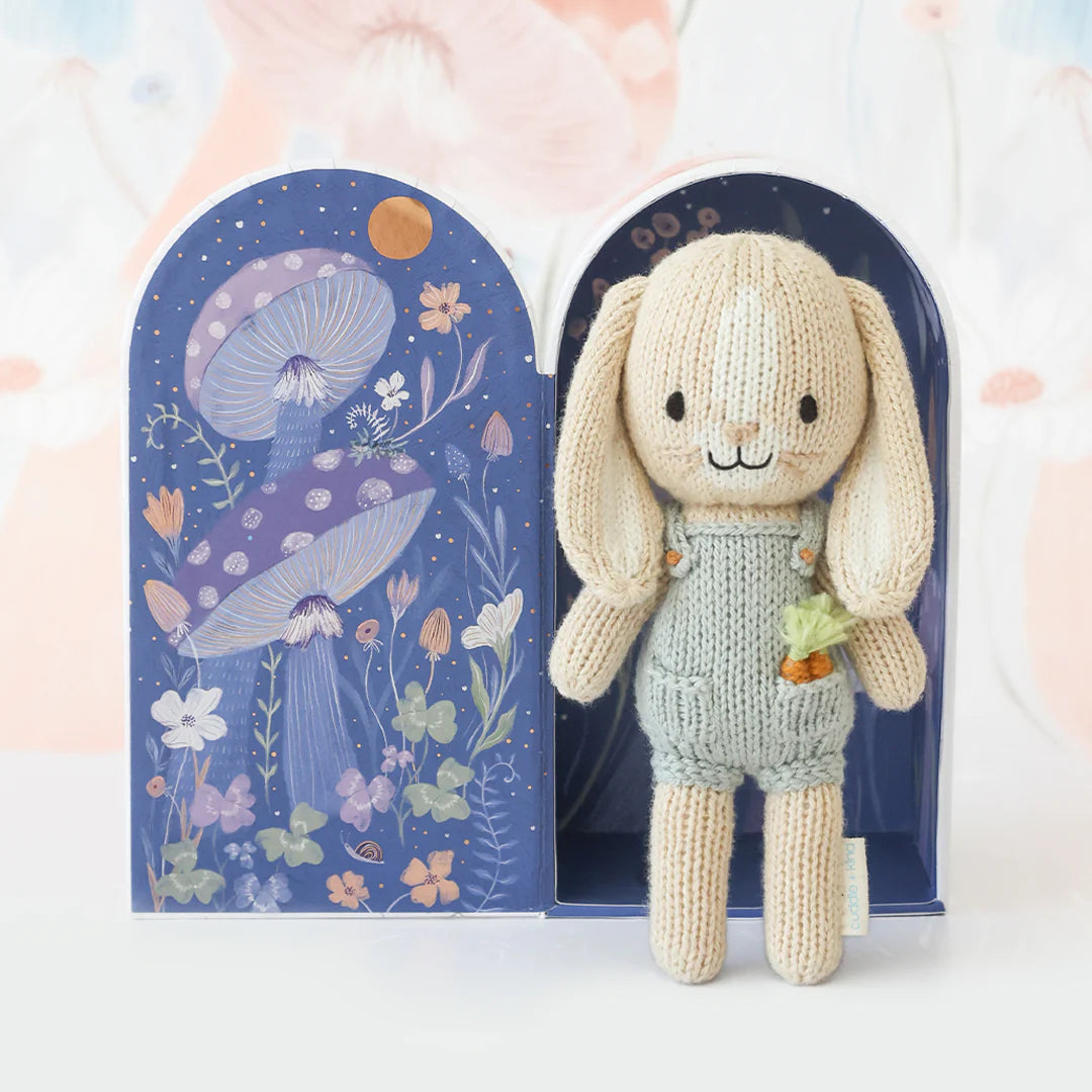 Meet Tiny Henry the Bunny—a hand-knit doll with long ears and a flower accessory from Cuddle + Kind's tiny collection, posed before an illustrated box of mushrooms and flowers. Ethically artisan-made and fair trade.