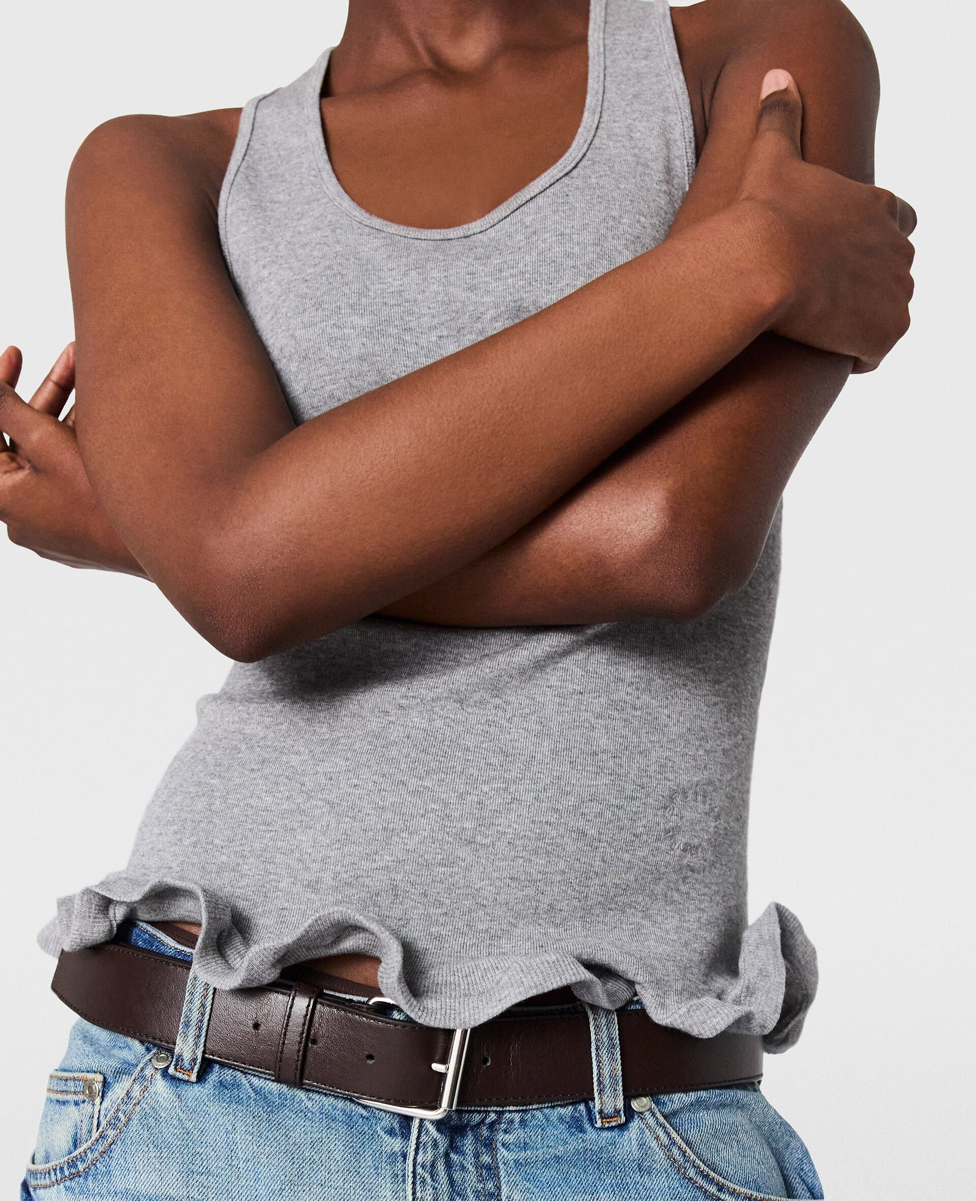 A person in the Stella McCartney Frilled Hem Rib Tank, blue jeans, and a brown belt stands with arms crossed.