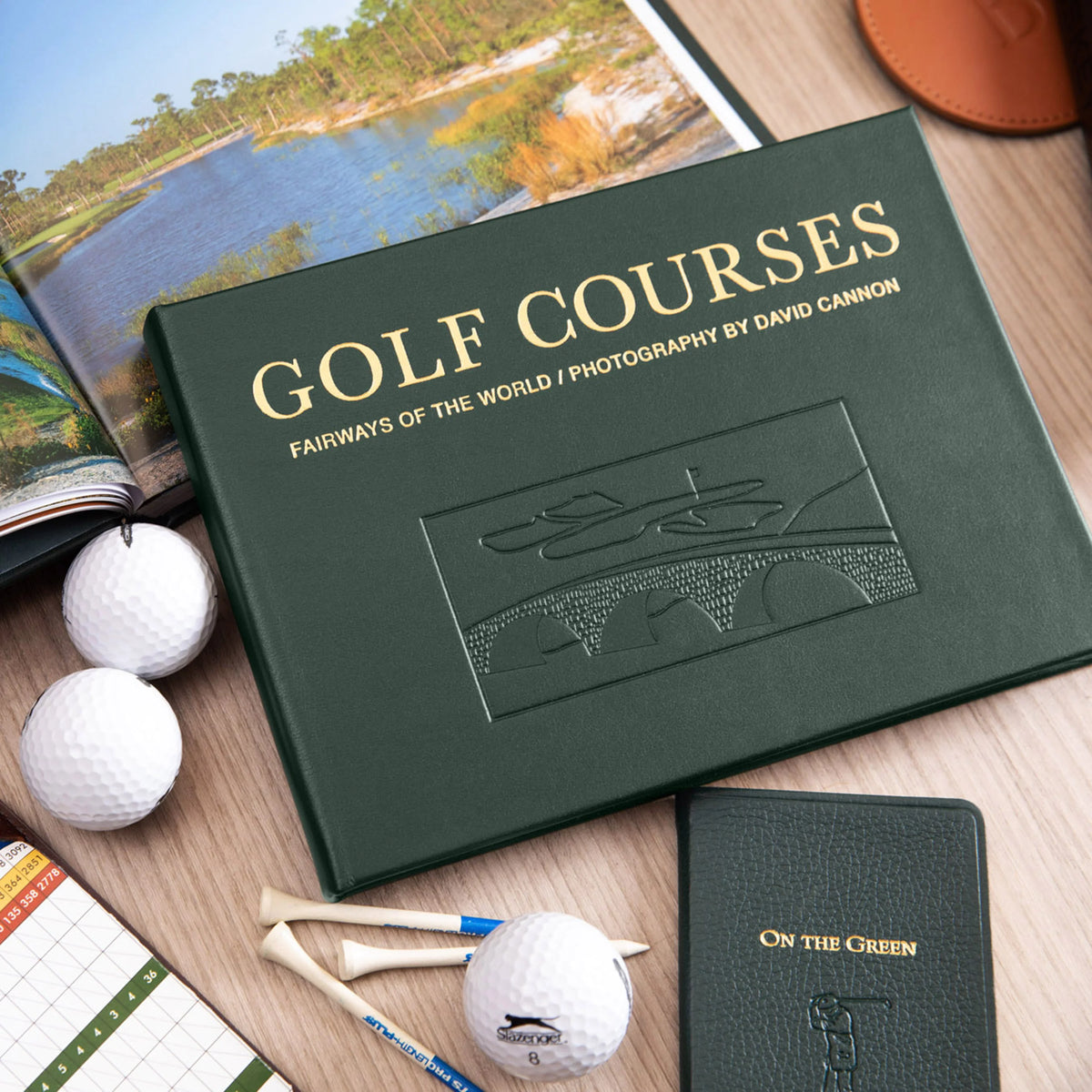 A green hardcover book titled “Golf Courses: Fairways of the World” featuring spectacular golf course photography is surrounded by golf balls, wooden tees, a scorecard, and a notebook on a wooden desk.