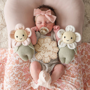 A newborn baby with a pacifier lies on a pink floral blanket, holding a handcrafted wooden “I’m Here” sign, flanked by two heirloom-quality Cuddle + Kind Blush Pink Daisy - Baby 7" flower-shaped plush toys.