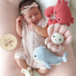 A newborn baby girl sleeps soundly on a pink blanket, surrounded by soft companions and knit toys—including Cuddle + Kind’s Baby Crab Coral, a cheerful flower, and a handcrafted heirloom baby animal whale—each lovingly made from cotton yarn. Nearby, a round wooden sign reads “It’s a girl.”.