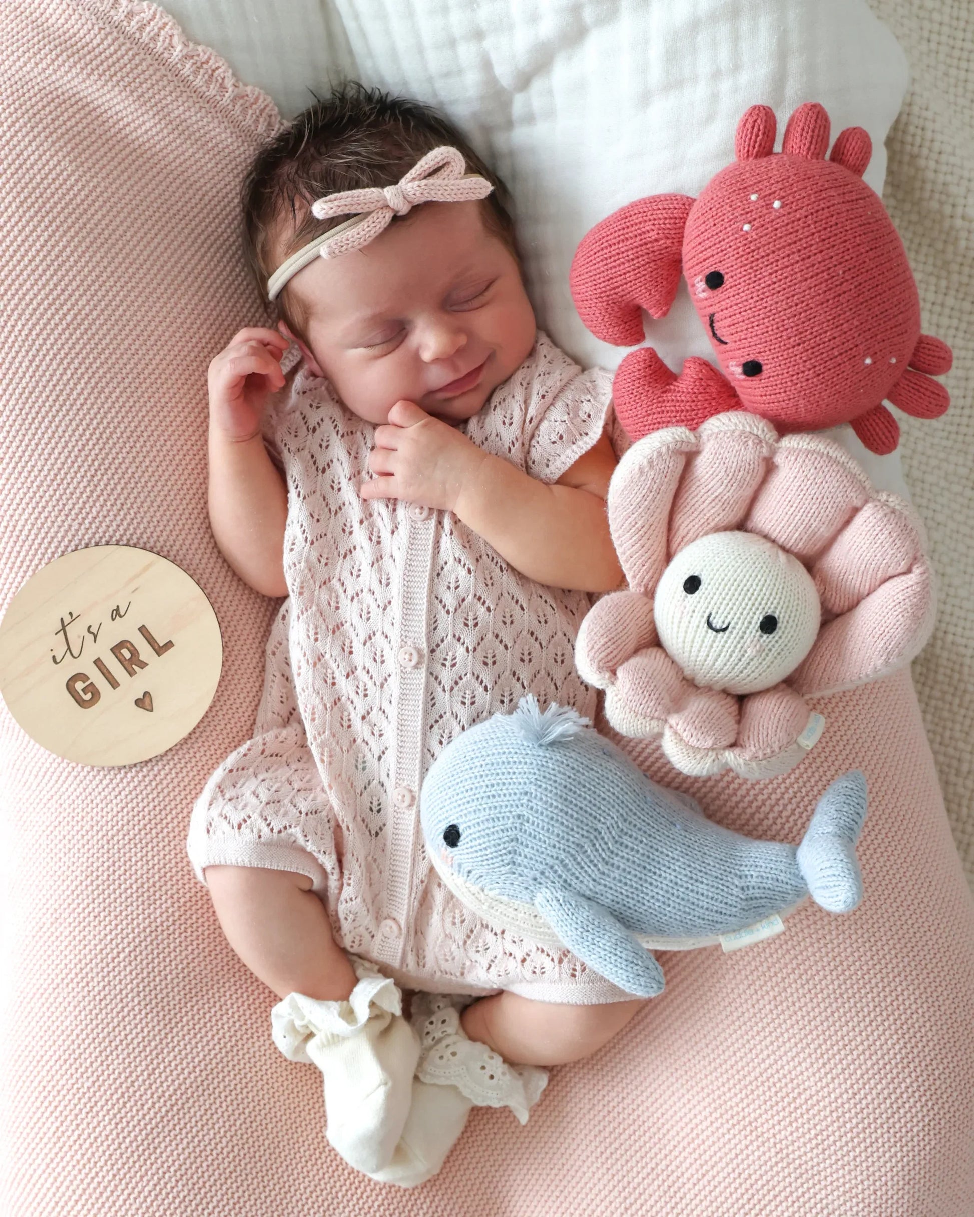 A newborn baby girl sleeps soundly on a pink blanket, surrounded by soft companions and knit toys—including Cuddle + Kind’s Baby Crab Coral, a cheerful flower, and a handcrafted heirloom baby animal whale—each lovingly made from cotton yarn. Nearby, a round wooden sign reads “It’s a girl.”.