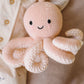 A Baby Octopus - Soft Coral Pink plush toy from Cuddle + Kind, crafted from soft Peruvian cotton yarn and featuring black eyes and a smiling mouth, rests against a cream-colored fabric background.