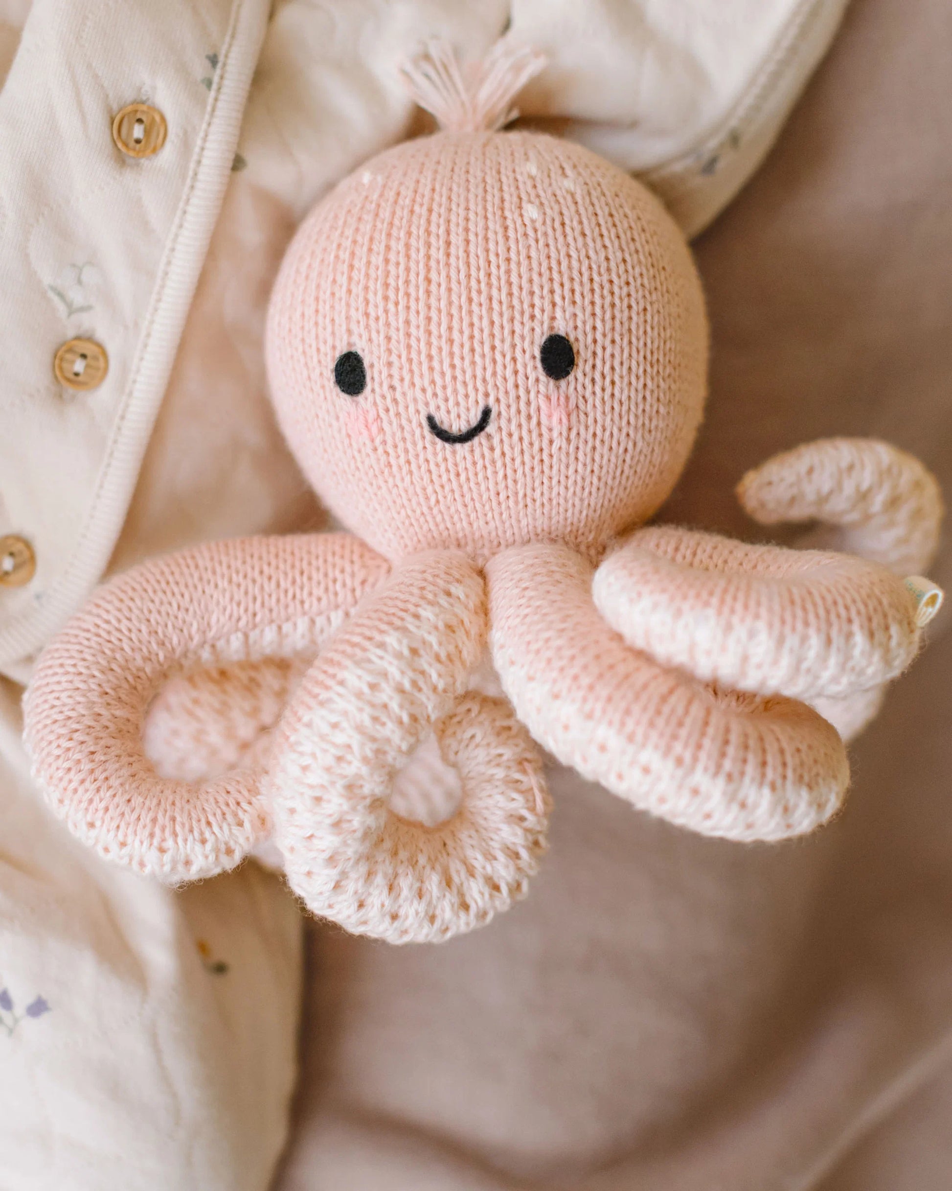 A Baby Octopus - Soft Coral Pink plush toy from Cuddle + Kind, crafted from soft Peruvian cotton yarn and featuring black eyes and a smiling mouth, rests against a cream-colored fabric background.