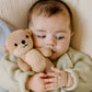 A baby in a light sweater lies on a blanket, holding a soft baby companion—a Cuddle + Kind Baby Otter crafted from heirloom-quality baby animal yarn and delicate handcrafted Peruvian cotton—close to their face.