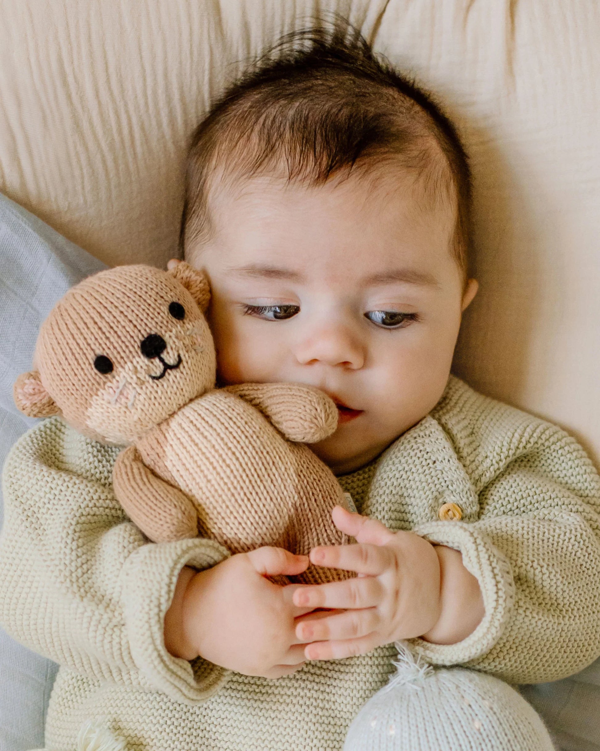A baby in a light sweater lies on a blanket, holding a soft baby companion—a Cuddle + Kind Baby Otter crafted from heirloom-quality baby animal yarn and delicate handcrafted Peruvian cotton—close to their face.
