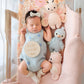 A newborn baby in a blue onesie and bow headband sleeps in a bassinet with a "hello world" sign, surrounded by three hand-knit heirloom-quality Cuddle + Kind baby animals, including the 7" Sky Blue Parrot, made from Peruvian cotton yarn on a pink blanket.