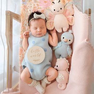 A newborn baby in a blue onesie and bow headband sleeps in a bassinet with a "hello world" sign, surrounded by three hand-knit heirloom-quality Cuddle + Kind baby animals, including the 7" Sky Blue Parrot, made from Peruvian cotton yarn on a pink blanket.