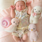 A newborn baby in a pastel knit outfit lies on a pink blanket, surrounded by three collectible stuffed toys: the Cuddle + Kind Snail - Baby 7", a hand-knit rabbit, and a bird—each baby animal crafted with heirloom-quality care.
