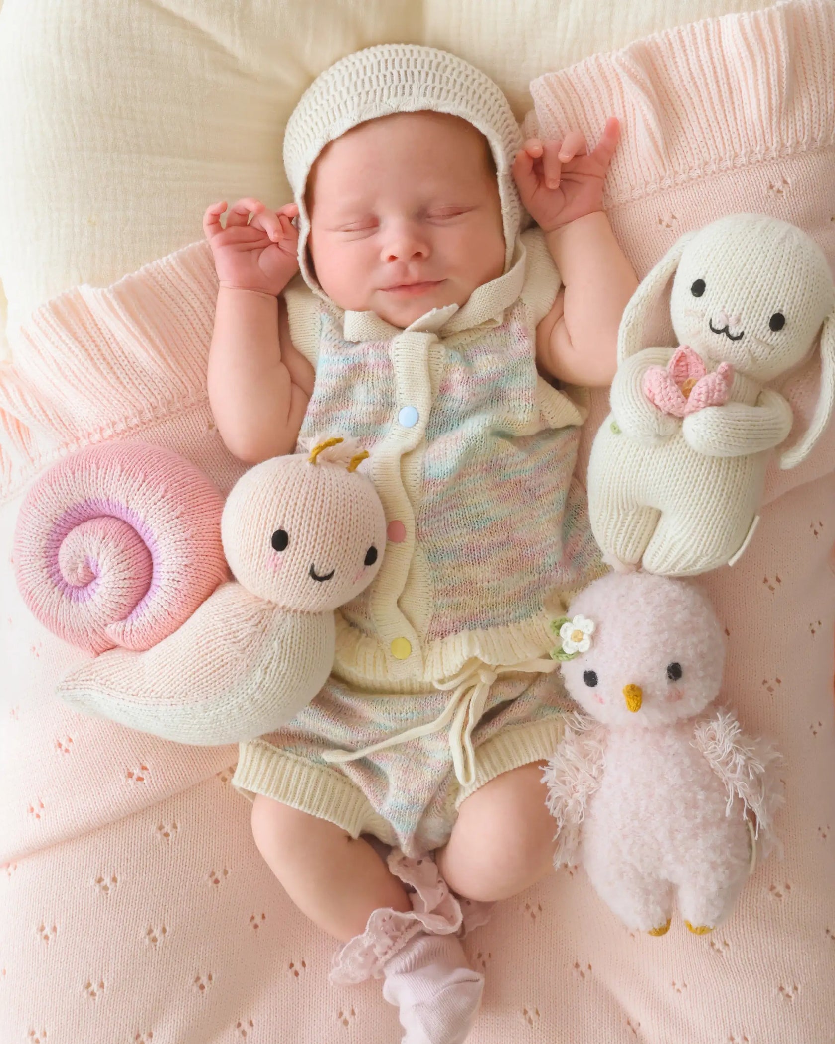 A newborn baby in a pastel knit outfit lies on a pink blanket, surrounded by three collectible stuffed toys: the Cuddle + Kind Snail - Baby 7", a hand-knit rabbit, and a bird—each baby animal crafted with heirloom-quality care.