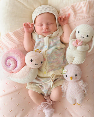 A newborn baby in a pastel knit outfit lies on a pink blanket, surrounded by three collectible stuffed toys: the Cuddle + Kind Snail - Baby 7", a hand-knit rabbit, and a bird—each baby animal crafted with heirloom-quality care.