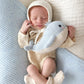 A newborn baby in a knit bonnet and outfit sleeps while hugging the Cuddle + Kind Baby Whale—a light blue, heirloom-quality companion lovingly handcrafted from Peruvian cotton—resting atop a soft white and light blue blanket.