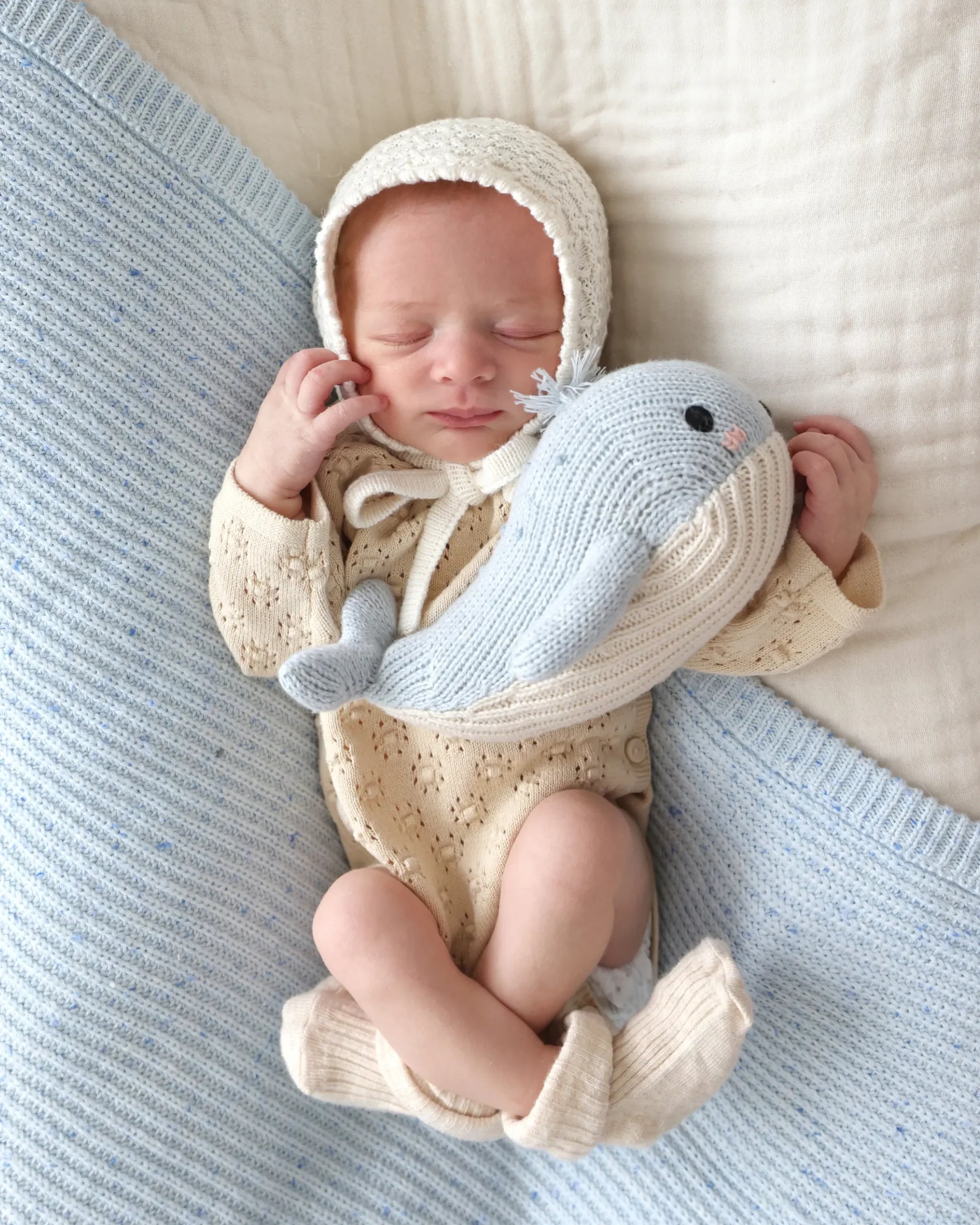 A newborn baby in a knit bonnet and outfit sleeps while hugging the Cuddle + Kind Baby Whale—a light blue, heirloom-quality companion lovingly handcrafted from Peruvian cotton—resting atop a soft white and light blue blanket.