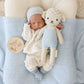 A newborn baby in a white outfit lies on a blue blanket, holding Cuddle + Kind's Benjamin the Lamb - Little 13", with a round sign reading "my first Easter" to the side—a charming gift for kids and the perfect keepsake for special moments.