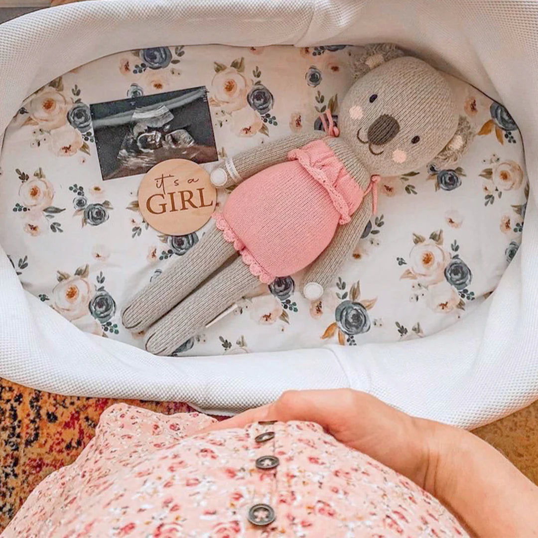 A hand rests on a floral-patterned baby bump above a crib with Claire the Koala - Little 13" by Cuddle + Kind, an “It’s a Girl” sign, and a sonogram photo—perfect for dreaming of treetop and treehouse adventures.